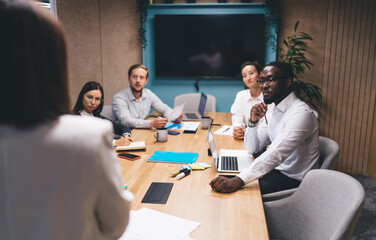 Concentrated young woman with glasses writing notes during tech-driven team strategy session, surrounded by digital tools and diverse coworkers in a smart office environment