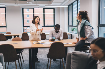 Asian woman and Black male colleague share ideas using a smartphone at a wooden table, symbolizing mobile technology use, digital collaboration, and smart teamwork in business context
