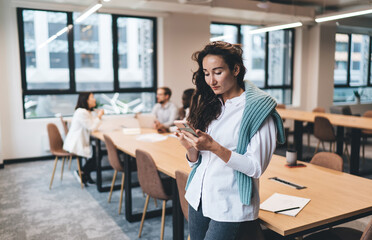 Confident young Caucasian businesswoman using smartphone for work tasks while standing in open office space, focused on mobile communication, digital productivity, or online research