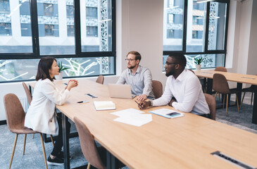 Three business professionals have a focused conversation at a table with laptops and documents, illustrating high-level planning, teamwork, and digital transformation in a modern office