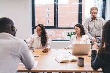 Startup team gathered around laptop and paper documents, analyzing charts and data during office planning session, actively engaging in strategy discussion in creative workspace