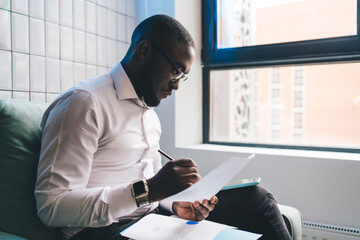 Young Black businessman in glasses reviewing documents in modern office lounge with natural light. Focused, professional mood, indoor workspace with city view