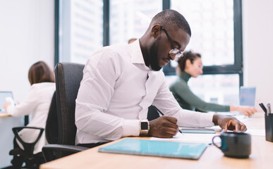 Focused African-American businessman writing at desk in modern office, surrounded by coworkers using laptops, working on reports and data analysis in tech-driven coworking space