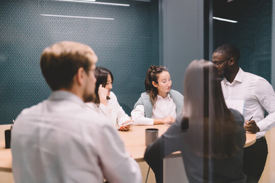 Diverse team having brainstorming session in glass meeting room, discussing ideas and digital tools around table, focused on collaborative tech project in startup workspace