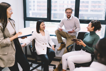 Office colleagues laughing and discussing project during informal meeting, standing and sitting around desk, showing teamwork, equality and positive work culture in startup workspace