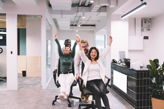 Multiethnic colleagues burst into laughter while racing chairs through office, capturing fun, unity, and emotional recharge in relaxed, engaging workplace environment - Powered by Adobe