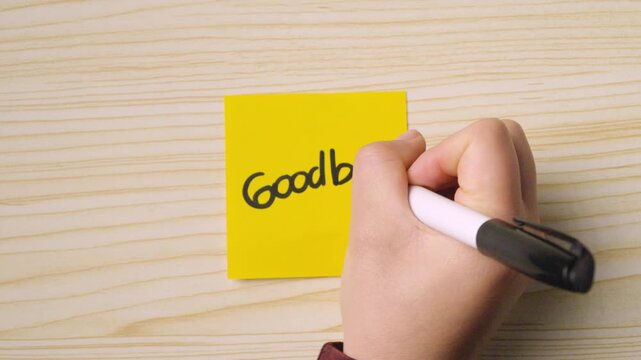 A hand holding a marker writing "Goodbye" with a smiley face on a yellow sticky note placed on a wooden surface.