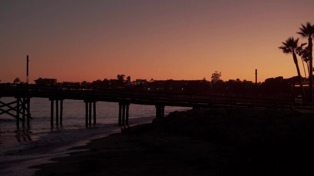 Silhouetted wooden pier with palm trees and city skyline at sunset