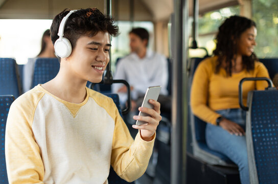 Laughing Asian guy using mobile phone sitting on city bus seat, happy male passenger in wireless headphones listening to music, watching video or movie, making online call, enjoying travel ride