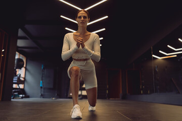 Young woman in white activewear holds deep lunge with arms centered, exuding mental focus, physical readiness, and elegance in minimalist gym space with striking overhead lights.
