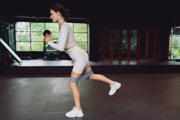 Slim fit woman performing leg kickback with elastic band, balancing on one foot with control, engaging glutes and core muscles for stability and strength enhancement.