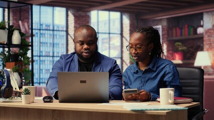 African American couple reviews taxes and finances at home office, using a calculator and laptop to track expenses, manage bills and plan their budget at home. Financial responsibility. Camera B.
