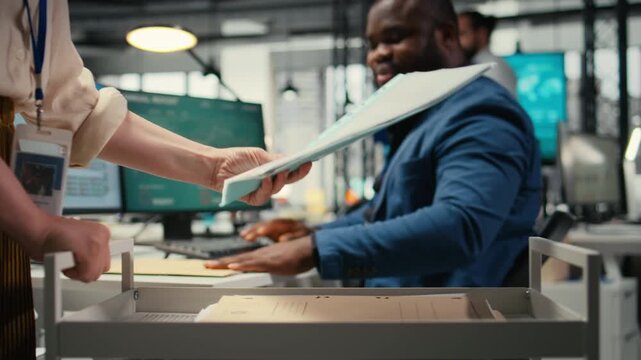 Professional archivist uses rolling trolley to transport documents and records, assisting the ceo and coworkers with efficient handling of critical paperwork in a modern workplace. Camera A.
