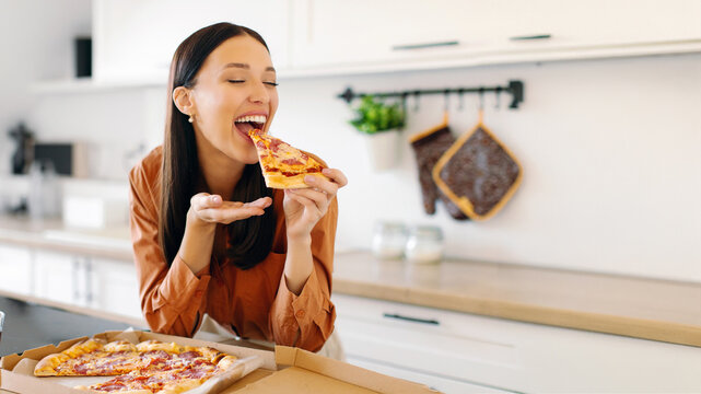 Excited young woman eating pizza, biting slice and enjoying its smell and taste, being satisfied with fast food delivery service. Lunch time at home - Powered by Adobe