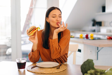 Young lady licking her finger while eating tasty hamburger, ordering takeaway food, enjoying delicious burger in kitchen at home