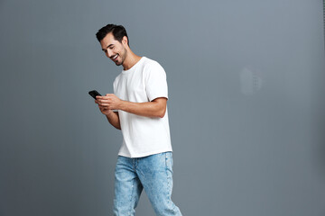 Full body smiling young man wearing white t-shirt and blue jeans, looking at his mobile phone with...