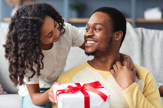 Adorable black girl greeting her smiling young father, hugging him and giving gift box, living room interior, copy space. Happy african american daddy and daughter celebrating Fathers day at home - Powered by Adobe