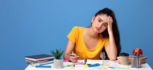 Tiresome Task. Portrait of exhausted frustrated young indian woman sitting at table, writing in notebook, resting head on hand. Sad female student tired of work, feeling headache, looking at camera