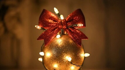 Ornament with red bow and lights A gold bauble adorned with a glittery red bow and wrapped in string lights illuminated against a warm neutral backdrop - Powered by Adobe
