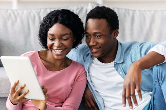 Smiling black lovers with digital tablet sitting on floor in living room, having idea to clean apartment, closeup. Happy african american young man and woman looking for house-keeping service - Powered by Adobe