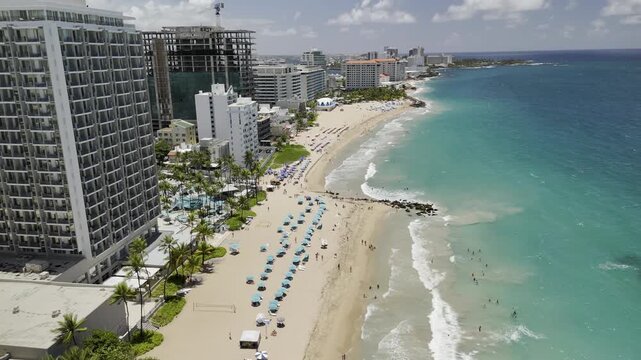 Drone flies west along Atlantic Beach covered in umbrellas as waves crash shore on sunny morning in San Juan, Puerto Rico