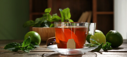 Glass cup of tasty hot tea, mint and limes on wooden table, closeup