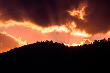 Sunlight bursts from behind volcanic peaks, casting golden rays across fluffy clouds and a deep...