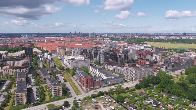 Aerial view of Copenhagen, Denmark, showcasing red roofed buildings, modern architecture, green spaces, and a clear blue sky with scattered clouds.