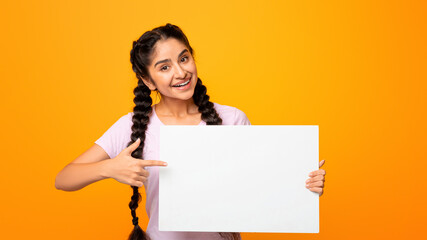 Place For Your Ad. Portrait of smiling indian lady holding empty blank board isolated on orange studio background. Happy woman standing with white square paper for template and pointing at it