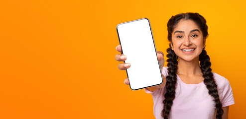 I Recommend. Positive young asian female showing mobile phone with black blank screen in hand, showing gadget close to camera. Smiling lady standing over yellow wall holding device for mock up