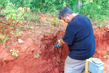 Naklejka premium Worker man uses jackhammer tools to dig remove roots from reddish soil while surrounded by greenery trees.