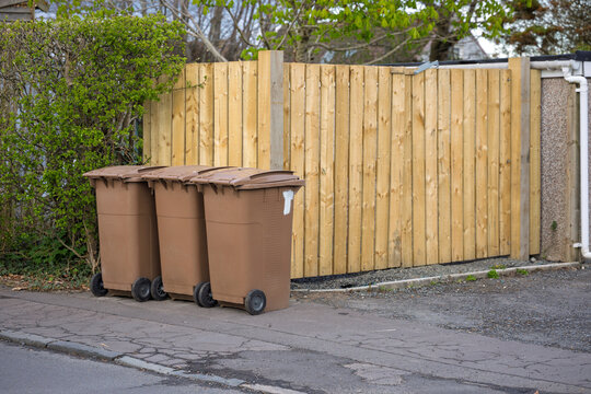Brown waste recycling wheelie bins on pavement beside wooden fence with green hedge
