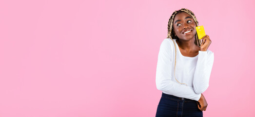 It's Shopping Time. Portrait of dreamy casual young African American woman holding credit card and looking away at copy space, thinking about sales and purchases, posing over pink studio background