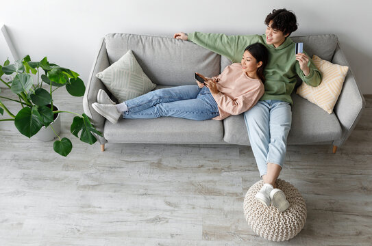 Happy Asian wife and husband sitting together on couch in living room, holding smartphone and credit card, shopping online, top view. Millennial couple using remote banking service at home, copy space - Powered by Adobe