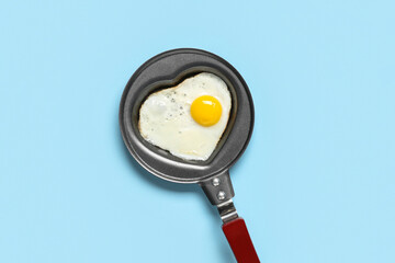 Frying pan with heart-shaped fried egg on blue background