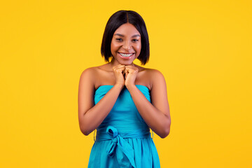 Sweet happy black lady in elegant blue summer dress putting hands together under her chin, smiling...