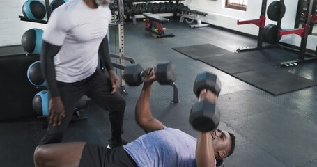 On trainer cue African American man pushing dumbbells on bench while trainer spotting for strength