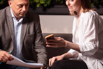 Two colleagues analyze data on a smartphone during a serious business meeting. Modern cozy office, greenery on the wall, documents and calculator on the table add realism. Preparation before the deal.