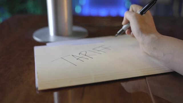 This video shows a close up, POV view of a hand writing "TARIFFS" on the front of a manilla folder on an office desk. 