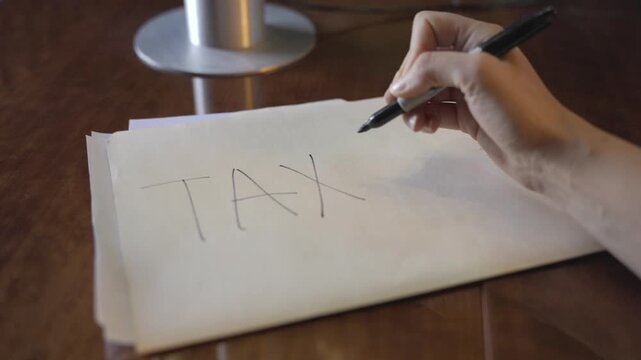 A close-up POV shot of a hand writing &ldquo;TAXES&rdquo; on the front of a manila folder placed on an office desk. The clip conveys themes of business, finance, and organization.