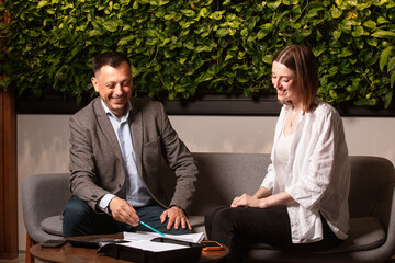 Two business partners laughing together while discussing documents in a modern office. Friendly atmosphere, green plant wall, teamwork. The director and his subordinate assistant are happy about deal.