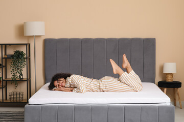 Young African-American woman with pillow lying on soft mattress in bedroom