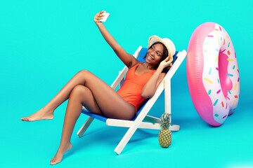 Gorgeous black woman taking selfie with smartphone in lounge chair over blue background, banner...
