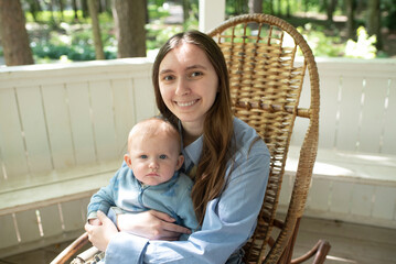 Fototapeta premium Young woman with long hair sits with baby child on rocking chair, in summer in gazebo, on sunny day. focus on the woman.
