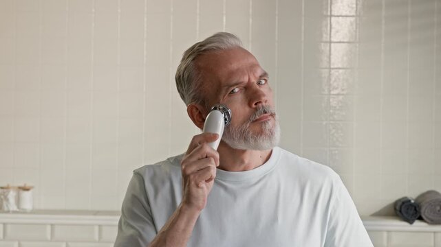 Bearded senior man looking at camera while standing in bathroom during morning hygiene and self care rituals and massaging his face with electric massager