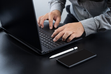 Businessman hands typing on laptop keyboard with smartphone and stylus pen on black desk workspace