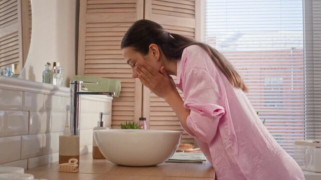 Side view of young brunette woman in pink shirt bending over sink while washing her face with cool water from tap, then taking soft towel and wiping clean skin