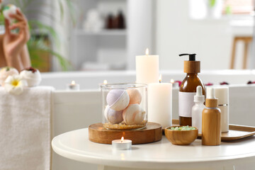 Bath bombs with cosmetic products and candles on coffee table against young woman in bathtub at...