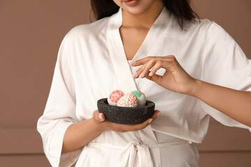 Pretty young woman in bathrobe holding bowl with bath bombs on brown background