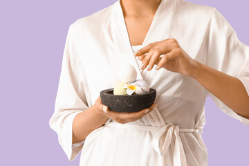 Pretty young woman in bathrobe holding bowl with bath bombs and plumeria flower on lilac background